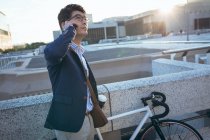 Азіатський бізнесмен говорить на смартфоні, тримаючи каву на своєму велосипеді на вулицях міста. Цифровий кочівник і про про про в міській концепції. — стокове фото