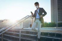 Азиатский бизнесмен в маске с помощью смартфона на городской улице. Цифровой кочевник в городе во время ковида 19 пандемической концепции. — стоковое фото