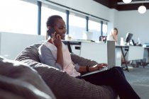 Lächelnde afrikanisch-amerikanische Geschäftsfrau, die im Sessel sitzt und bei der Arbeit mit dem Smartphone spricht. unabhängiges kreatives Geschäft in einem modernen Büro. — Stockfoto