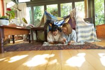 Улыбающаяся азиатка со своей дочерью, использующая планшет, лежащий под палаткой в гостиной. в доме в изоляции во время карантинной изоляции. — стоковое фото