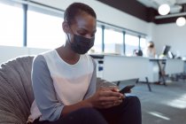 Mujer de negocios afroamericana sonriente sentada en un sillón, con máscara facial, usando un teléfono inteligente. negocio creativo independiente en una oficina moderna durante coronavirus covid 19 pandemia. - foto de stock
