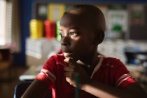 Niño afroamericano mirando por la ventana mientras está sentado en su escritorio en clase en la escuela primaria. escuela y concepto de educación - foto de stock