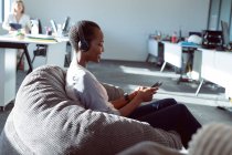 Empresária afro-americana sorridente sentada em poltrona, usando fones de ouvido, usando smartphone. negócio criativo independente em um escritório moderno. — Fotografia de Stock