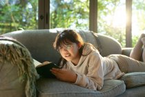 Lächelndes asiatisches Mädchen mit Brille, das ein Buch liest und auf dem Sofa liegt. Zuhause in Isolation während der Quarantäne. — Stockfoto