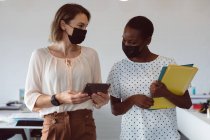 Zwei unterschiedliche Geschäftsfrauen mit Mundschutz, Tablet und Dokumenten in der Hand, unterhalten sich. Selbstständiges kreatives Geschäft in einem modernen Büro während der Coronavirus-Pandemie 19. — Stockfoto