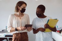 Duas mulheres de negócios diversas usando máscaras faciais, segurando tablet e documentos, conversando. negócio criativo independente em um escritório moderno durante coronavírus covid 19 pandemia. — Fotografia de Stock