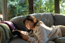 Menina asiática lendo um livro e deitado no sofá. em casa em isolamento durante o confinamento de quarentena. — Fotografia de Stock