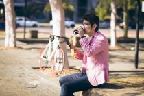 Asiatischer Geschäftsmann, der mit seinem Smartphone Kaffee zum Mitnehmen trinkt, sitzt in der Stadt an der Wand. Digitaler Nomade im Stadtkonzept unterwegs. — Stockfoto