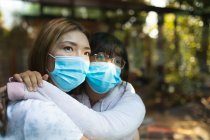Traurige asiatische Frau und ihre Tochter umarmen sich mit Mundschutz und schauen aus dem Fenster. Zuhause in Isolation während Covid 19 Pandemie und Quarantäne Lockdown. — Stockfoto
