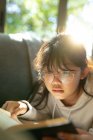 Menina asiática em óculos lendo um livro e deitado no sofá. em casa em isolamento durante o confinamento de quarentena. — Fotografia de Stock