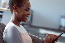 Femme d'affaires afro-américaine souriante assise dans un fauteuil, à l'aide d'un smartphone. entreprise créative indépendante dans un bureau moderne. — Photo de stock
