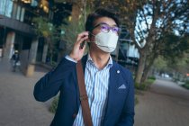 Азиатский бизнесмен в маске с помощью смартфона на городской улице. Цифровой кочевник в городе во время ковида 19 пандемической концепции. — стоковое фото
