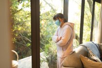 Sad asian girl in glasses wearing face mask and looking out of window. at home in isolation during covid 19 pandemic and quarantine lockdown. — Stock Photo