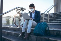 Homme d'affaires asiatique portant un masque facial à l'aide d'un ordinateur portable assis sur les marches dans la rue de la ville. numérique nomade dehors et environ dans la ville pendant covid 19 concept pandémique. — Photo de stock