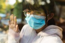 Triste menina asiática em óculos usando máscara facial e olhando para fora da janela. em casa, isoladamente, durante o bloqueio da pandemia e quarentena do covid 19. — Fotografia de Stock