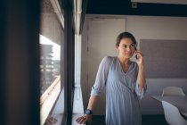 Kaukasische Geschäftsfrau, die am Fenster steht und bei der Arbeit mit dem Smartphone spricht. unabhängiges kreatives Geschäft in einem modernen Büro. — Stockfoto
