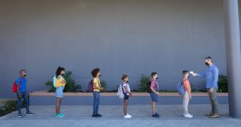 Professor caucasiano do sexo masculino medindo a temperatura de um grupo de estudantes em pé em uma fila na escola. higiene e distanciamento social na escola durante a pandemia de 19 anos — Fotografia de Stock