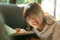 Chica asiática sonriente en gafas leyendo un libro y acostada en un sofá. en casa en aislamiento durante el bloqueo de cuarentena. - foto de stock