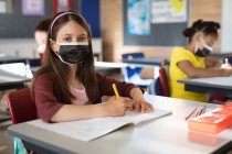 Retrato de menina caucasiana usando máscara facial sentada em sua mesa na classe na escola primária. higiene e distanciamento social na escola durante a pandemia de 19 anos — Fotografia de Stock