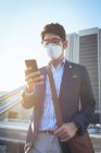 Asiatischer Geschäftsmann mit Gesichtsmaske und Smartphone auf der Straße der Stadt. digitaler Nomade während des Covid 19 Pandemiekonzepts in der Stadt unterwegs. — Stockfoto