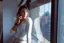 Kaukasische Geschäftsfrau, die am Fenster steht und bei der Arbeit mit dem Smartphone spricht. unabhängiges kreatives Geschäft in einem modernen Büro — Stockfoto