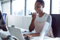 African american businesswoman sitting at desk, talking by smartphone, smiling. independent creative business at a modern office. — Stock Photo