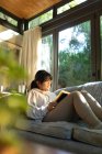 Усміхнена азіатська дівчина в окулярах читає книгу і сидить на дивані. вдома в ізоляції під час карантину . — стокове фото