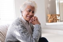Retrato de una mujer mayor de raza mixta sentada en un sofá mirando a la cámara y sonriendo. permanecer en casa aislado durante el bloqueo de cuarentena. - foto de stock