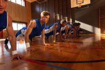 Equipo de baloncesto masculino diverso que usa ropa deportiva azul y hace flexiones. baloncesto, entrenamiento deportivo en una cancha cubierta. - foto de stock