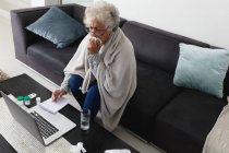 Femme âgée de race mixte assis sur le canapé faire appel vidéo à l'aide d'un ordinateur portable. rester à la maison dans l'isolement pendant le confinement en quarantaine. — Photo de stock