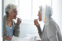 Donna anziana di razza mista che guarda il suo riflesso nello specchio con indosso il rossetto. stare a casa in isolamento durante la quarantena. — Foto stock