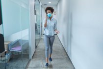 Donna d'affari di razza mista che indossa maschera facciale e parla su smartphone. lavoro in un ufficio moderno durante covid 19 coronavirus pandemia. — Foto stock