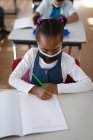 Ragazza afroamericana indossando maschera viso studiare mentre seduto sulla scrivania in classe a scuola. igiene e distanza sociale a scuola durante la covd 19 pandemia — Foto stock