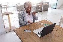 Mulher sênior de raça mista sentada à mesa usando laptop e tomando medicamentos. ficar em casa em isolamento durante o confinamento de quarentena. — Fotografia de Stock