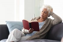 Femme âgée de race mixte assis sur le canapé livre de lecture. rester à la maison dans l'isolement pendant le confinement en quarantaine. — Photo de stock