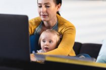 Madre caucasica che tiene in braccio il suo bambino usando il portatile mentre lavora da casa. maternità, amore e concetto di cura del bambino — Foto stock