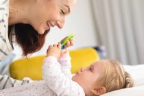 Madre caucásica sonriendo mientras su bebé juega con un juguete mientras está acostado en la cama en casa. maternidad, amor y cuidado del bebé - foto de stock