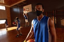 Porträt eines männlichen Basketballspielers gemischter Rasse mit Gesichtsmaske und Team im Hintergrund. Basketball, Sporttraining auf einem überdachten Platz während der Coronavirus-Pandemie 19. — Stockfoto