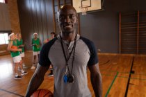Portrait de l'entraîneur afro-américain de basket-ball masculin avec l'équipe en arrière-plan. basket-ball, entraînement sportif sur un terrain intérieur. — Photo de stock