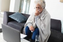 Mulher sênior de raça mista sentado no sofá fazendo chamada de vídeo usando laptop. ficar em casa em isolamento durante o confinamento de quarentena. — Fotografia de Stock