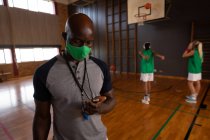 Entrenador de baloncesto masculino afroamericano con máscara facial con equipo de fondo. baloncesto, entrenamiento deportivo en una cancha de interior durante la pandemia de coronavirus covid 19. - foto de stock