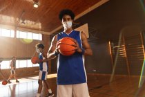 Retrato de mestiço jogador de basquete masculino vestindo máscara facial com a equipe em segundo plano. basquete, treinamento esportivo em um tribunal interno durante coronavírus covid 19 pandemia. — Fotografia de Stock