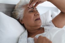 Gemischte Rasse Seniorin liegt im Bett und hält ihren Kopf in Gedanken. Isolationshaft während der Quarantäne. — Stockfoto