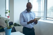 Uomo d'affari afroamericano in piedi accanto alla finestra, sorridente e utilizzando tablet. lavorare in un'attività creativa indipendente. — Foto stock
