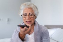 Donna anziana di razza mista seduta sul letto a parlare su smartphone. stare a casa in isolamento durante la quarantena. — Foto stock