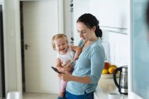 Mãe branca segurando seu bebê usando smartphone na cozinha em casa. maternidade, amor e conceito de cuidado do bebê — Fotografia de Stock
