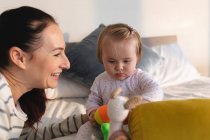 Mãe branca e seu bebê brincando com um brinquedo enquanto estão sentados na cama em casa. maternidade, amor e conceito de cuidado do bebê — Fotografia de Stock