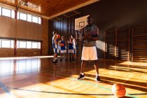 Entrenador de baloncesto masculino afroamericano con equipo de fondo. baloncesto, entrenamiento deportivo en una cancha cubierta durante. - foto de stock