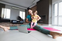 Кавказька мати тримає своє немовля, виконуючи розтягнуті вправи, дивлячись на ноутбук удома. материнство, любов і догляд дітей — стокове фото