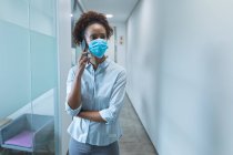 Mixed race businesswoman wearing face mask and talking on smartphone. work at a modern office during covid 19 coronavirus pandemic. — Stock Photo